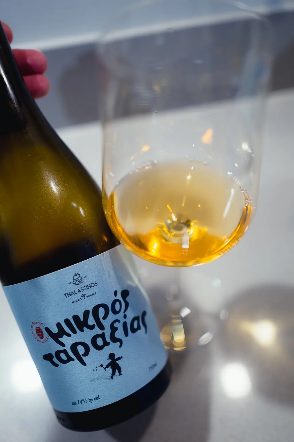 Hand houdt fles Mikros Taraxias vast naast glas amberkleurige wijn van bovenaf