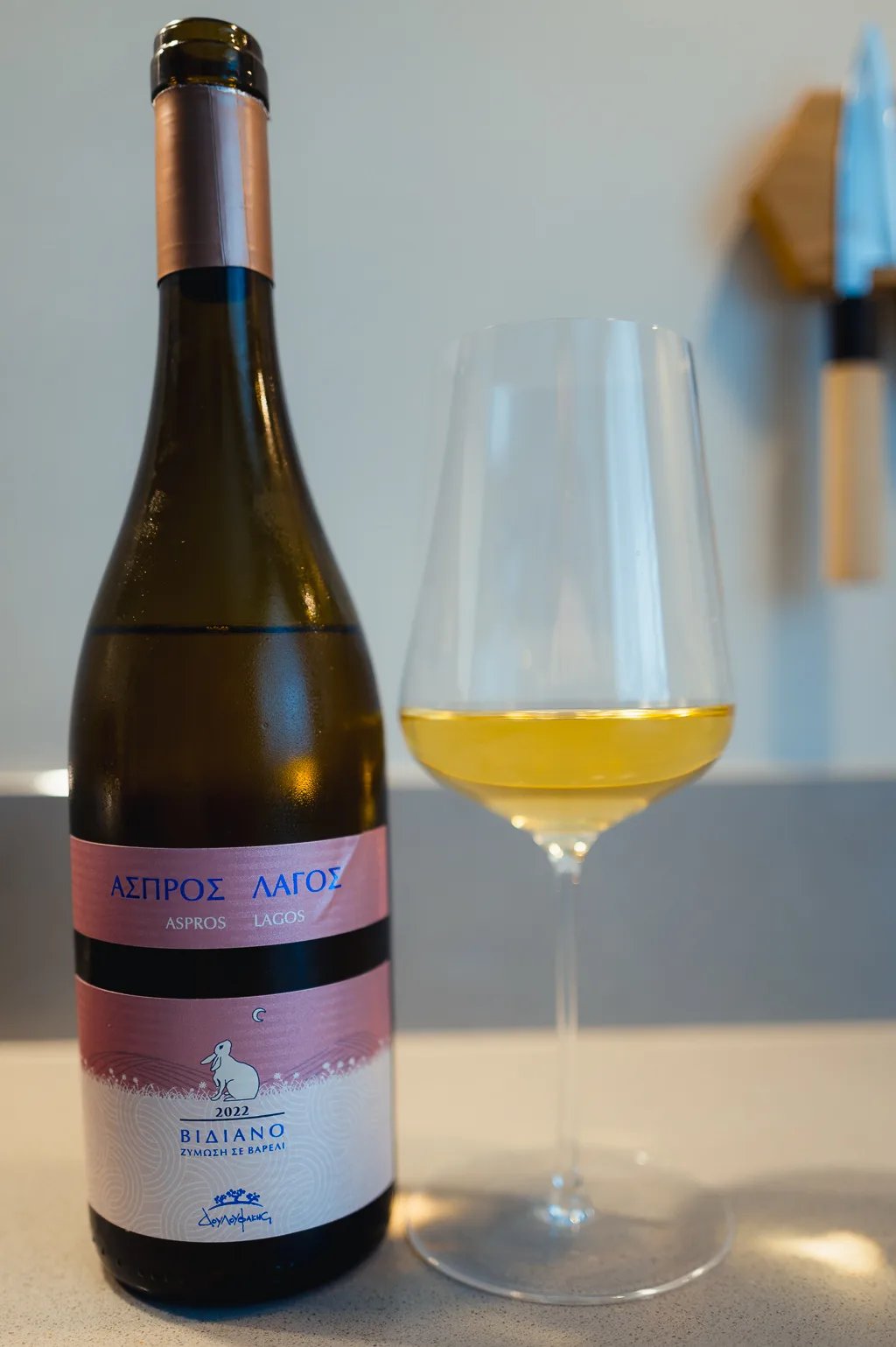 Fles Aspros Lagos Vidiano met roze etiket naast glas lichtgele Kretenzische wijn