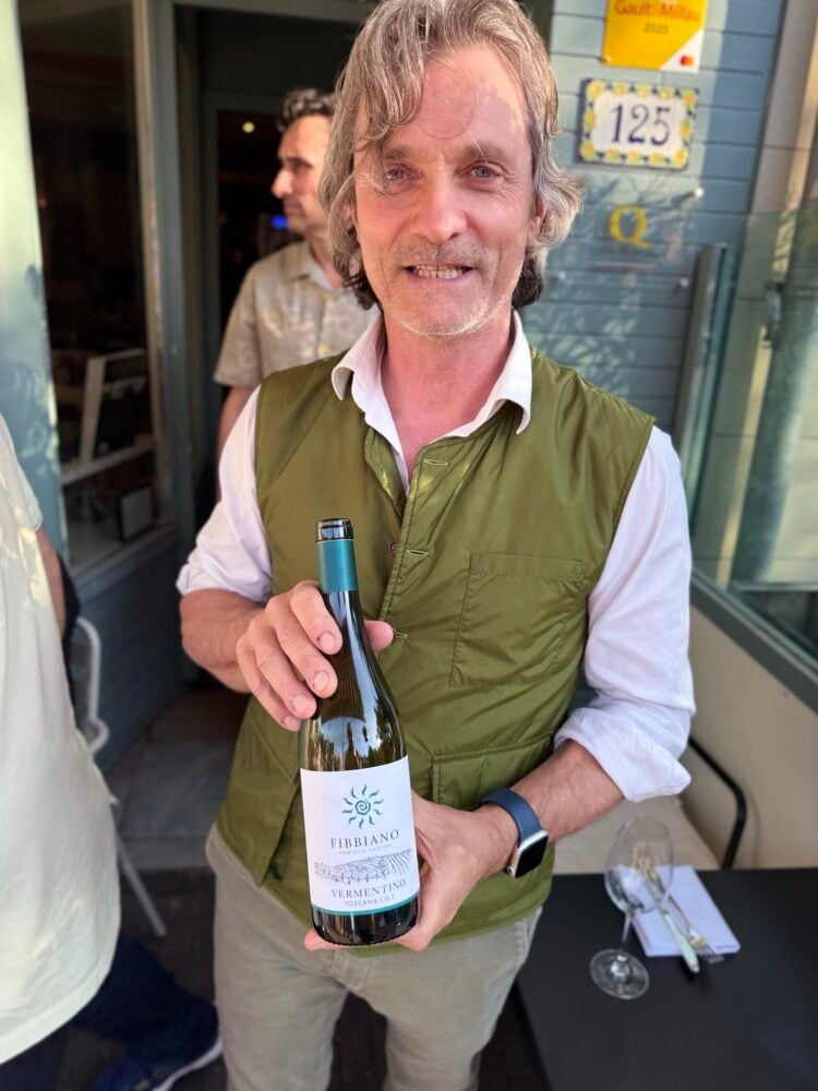 Glimlachende man in groen bodywarmer houdt fles Fibbiano Vermentino Toscana voor een gevel
