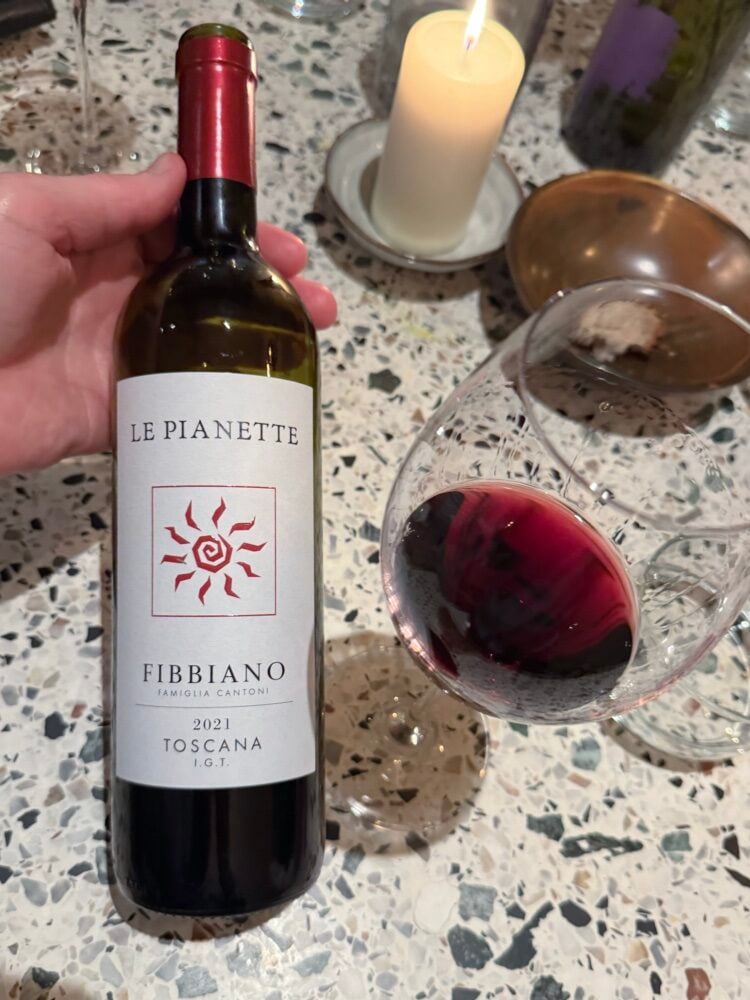 Hand houdt fles Fibbiano Le Pianette Toscana 2021 vast naast glas rode wijn bij kaarslicht