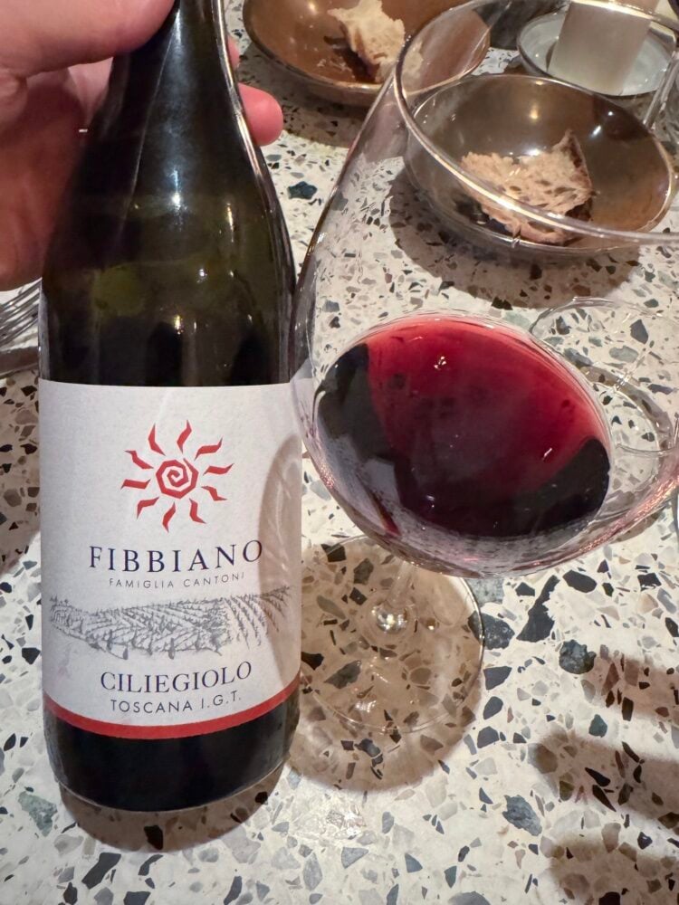 Hand houdt fles Fibbiano Ciliegiolo Toscana vast naast glas rode wijn op een terrazzo tafel