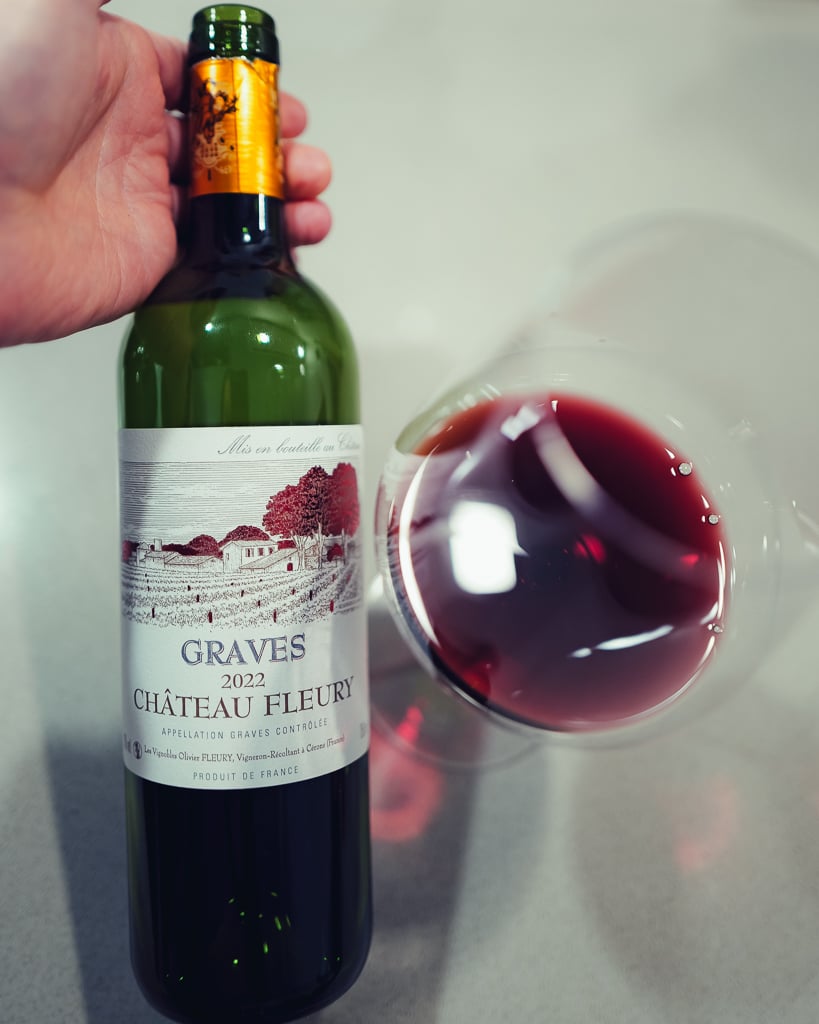 Hand houdt een fles Graves 2022 Château Fleury vast naast een liggend glas rode wijn