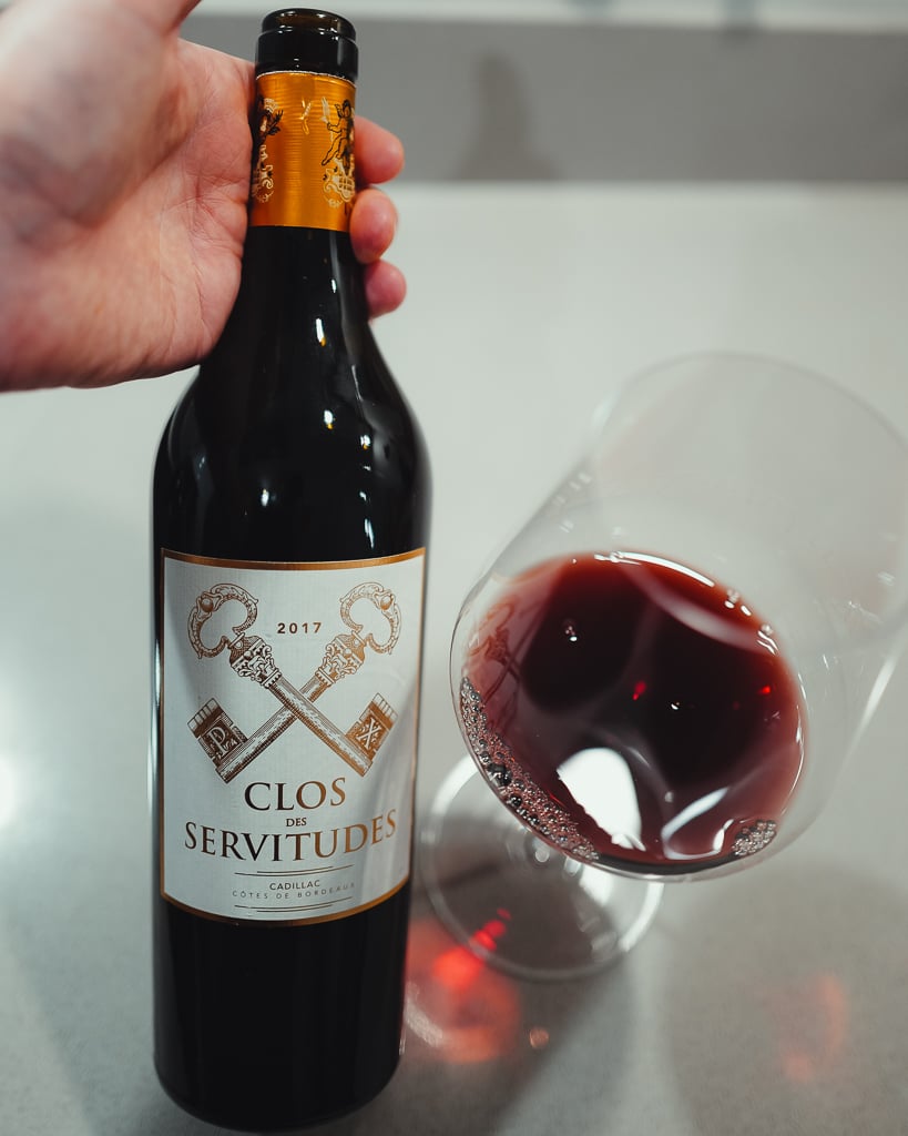Hand houdt een fles Château Gravelines 2023 vast naast een liggend leeg wijnglas