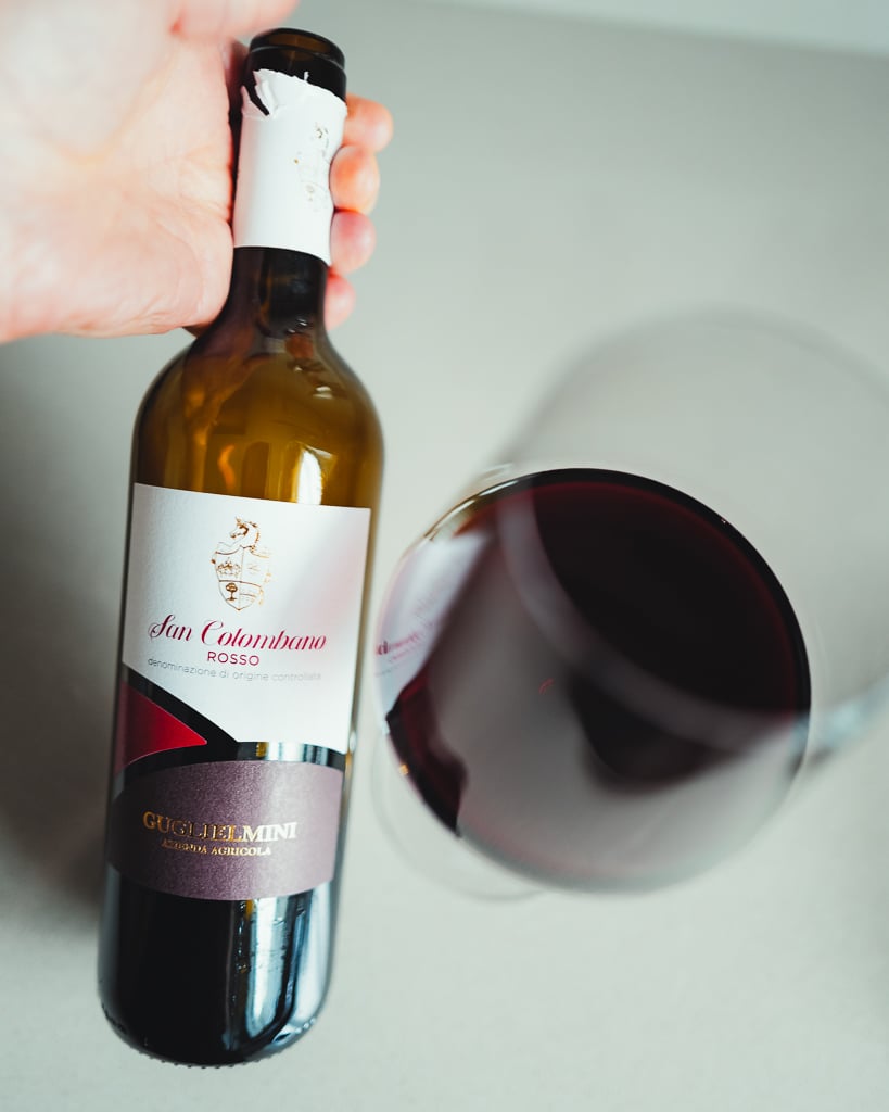 Hand houdt een fles San Colombano Rosso van Guglielmini vast naast een liggend glas rode wijn