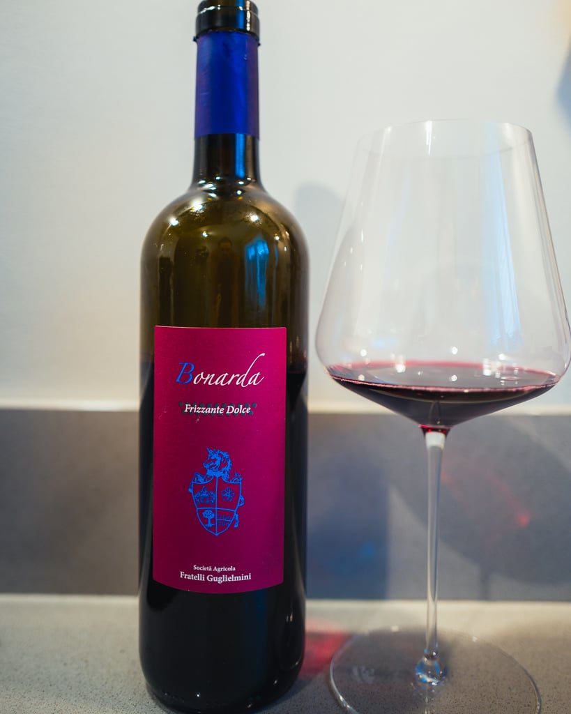 Fles Bonarda Frizzante Dolce van Guglielmini met roze etiket naast een glas rode wijn