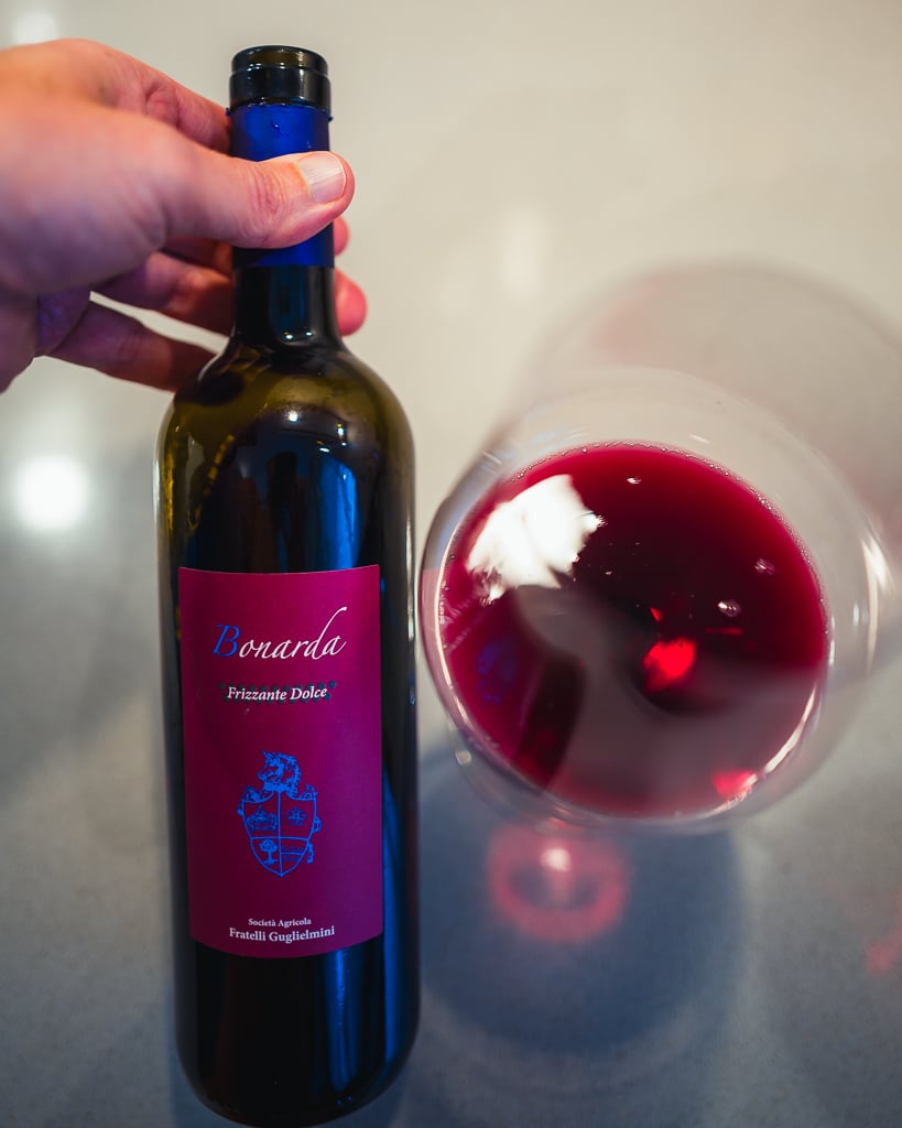 Hand houdt een fles Bonarda Frizzante Dolce vast naast een liggend glas felrode wijn