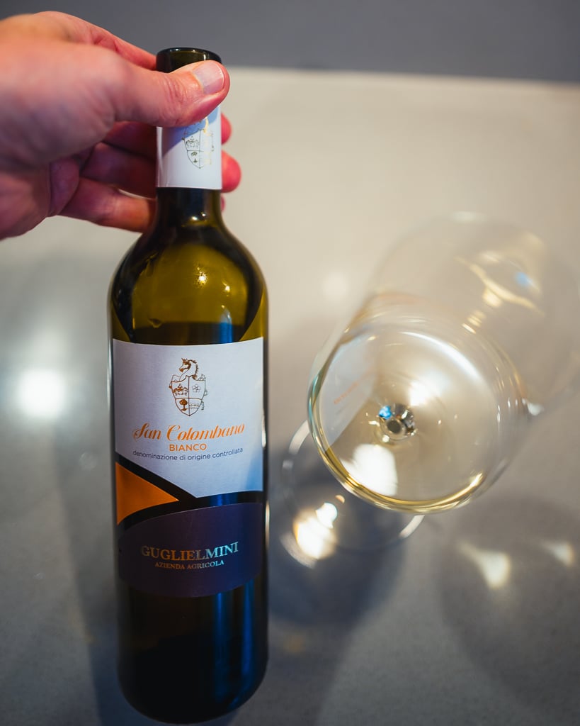 Hand houdt een fles San Colombano Bianco van Guglielmini vast naast een liggend glas witte wijn