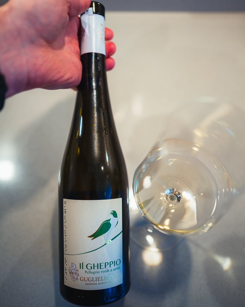 Hand houdt een fles Il Gheppio van Guglielmini vast naast een liggend glas witte wijn