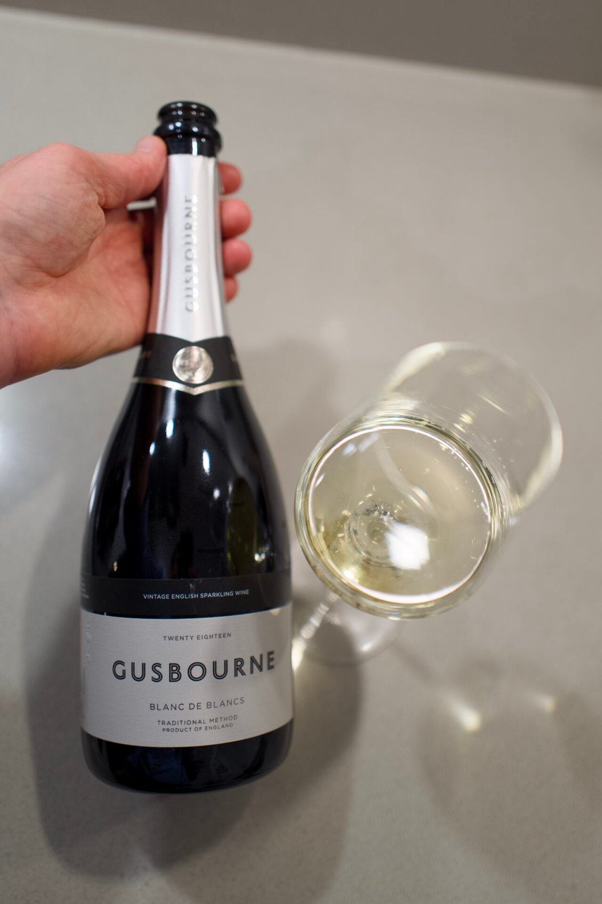 Hand houdt een fles Gusbourne Blanc de Blancs 2018 vast naast een liggend glas witte wijn