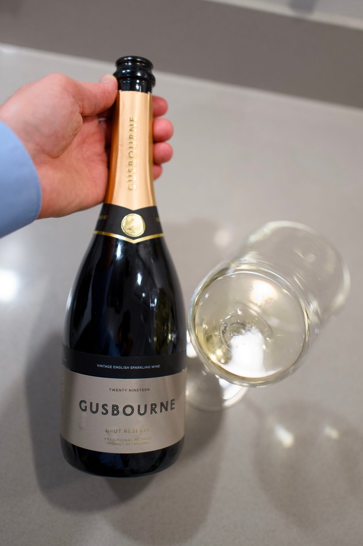 Hand houdt een fles Gusbourne Brut Reserve 2019 vast naast een liggend glas witte wijn