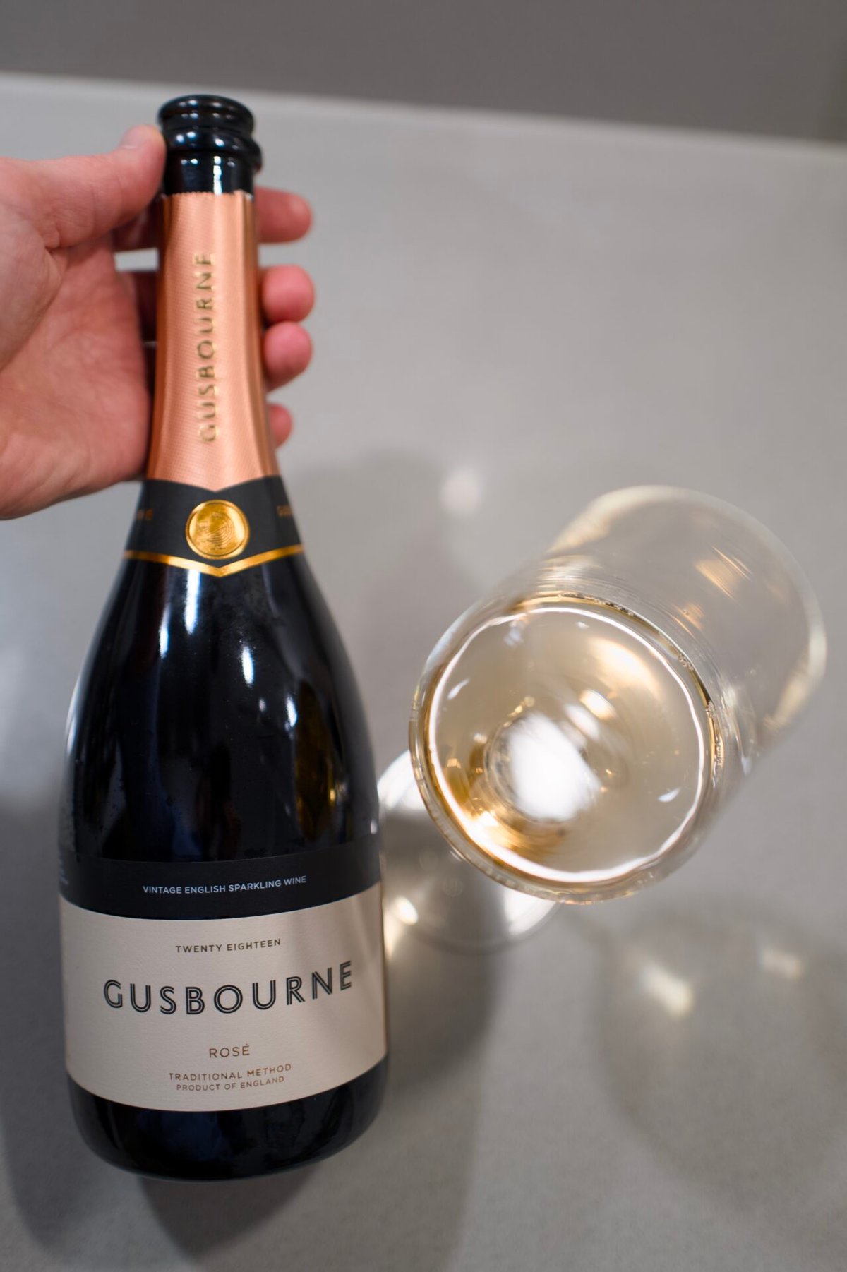 Hand houdt een fles Gusbourne Rosé 2018 vast naast een liggend glas wijn