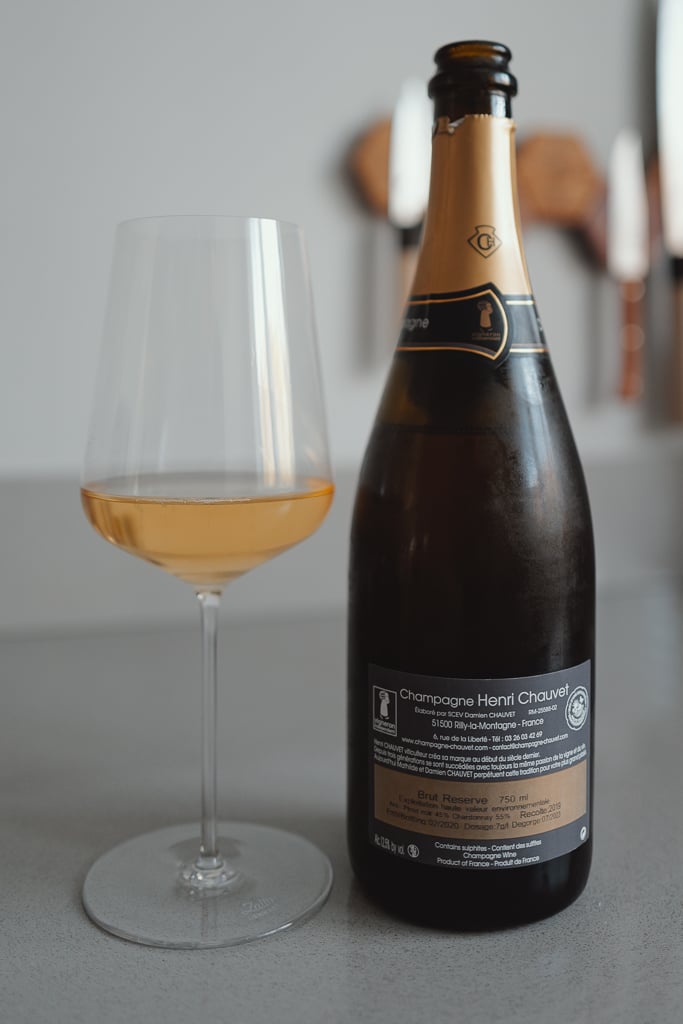 Glas amberkleurige champagne naast een fles met achteretiket van Champagne Henri Chauvet Brut Réserve