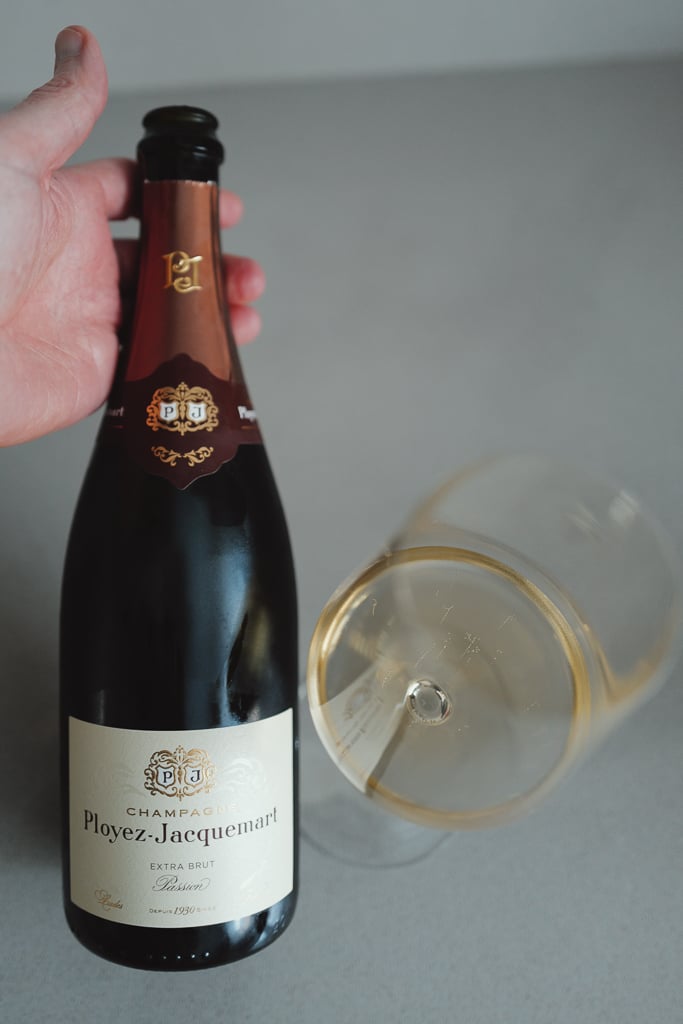 Hand houdt een fles Champagne Ployez-Jacquemart Extra Brut Passion vast naast een liggend glas champagne