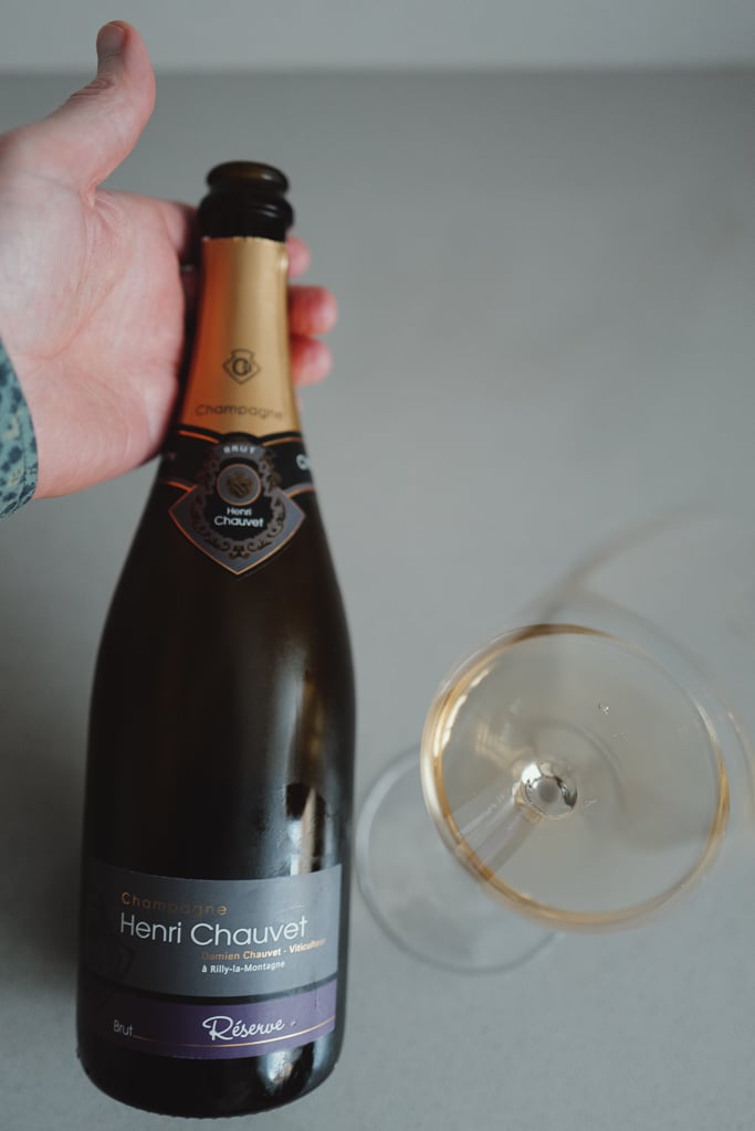 Hand houdt een fles Champagne Henri Chauvet Brut Réserve vast naast een liggend glas champagne
