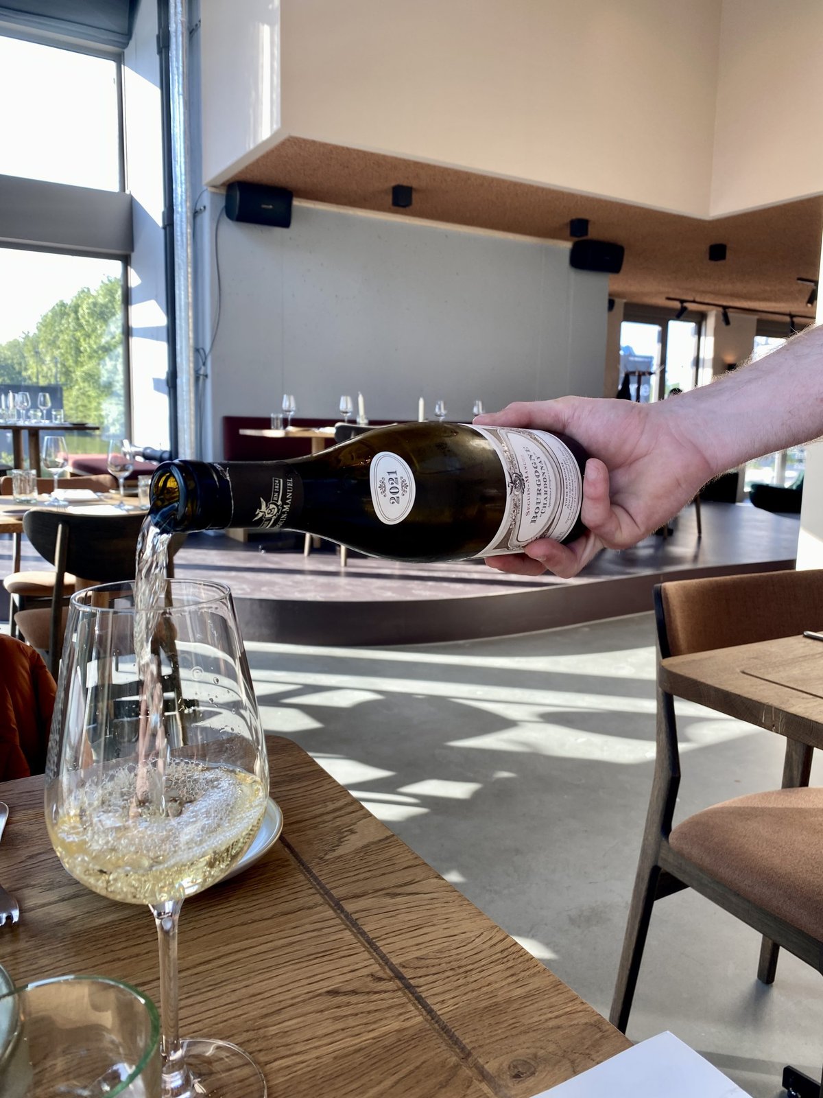 Hand schenkt mousserende wijn in een glas aan een houten tafel in een zonnig restaurant