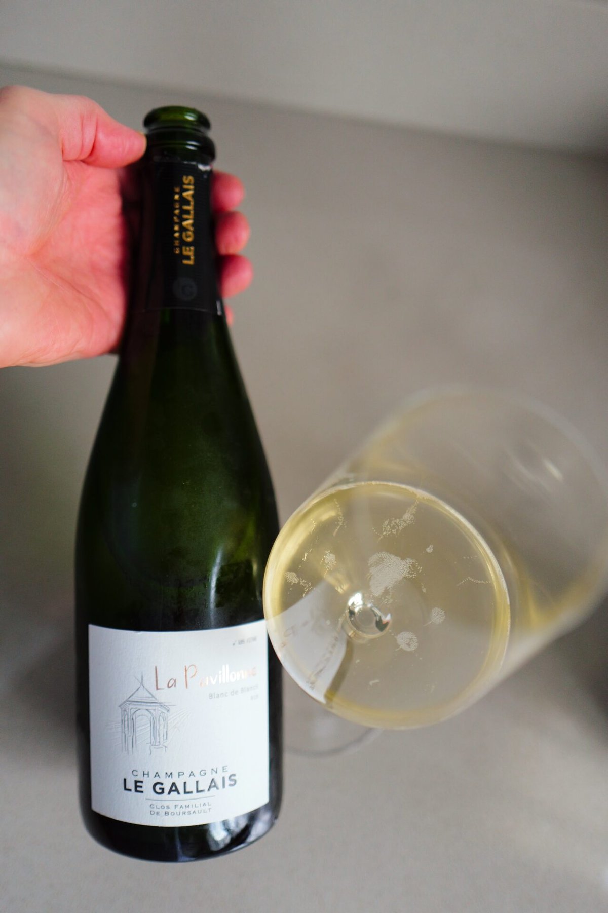 Hand houdt de La Pavillonne champagnefles vast naast een liggend glas met lichte champagne