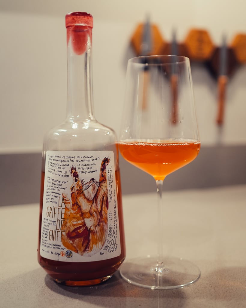 Fles La Griffe de Gniff oranjewijn met kat-illustratie op het etiket naast een glas amberkleurige wijn