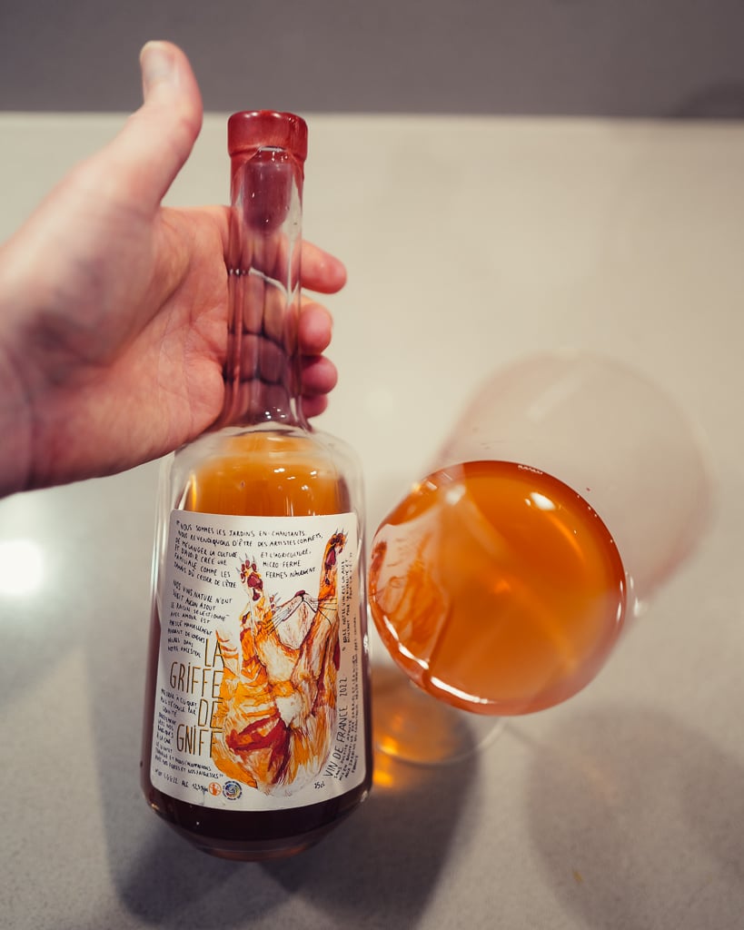 Hand houdt de fles La Griffe de Gniff oranjewijn vast naast een liggend glas amberkleurige wijn