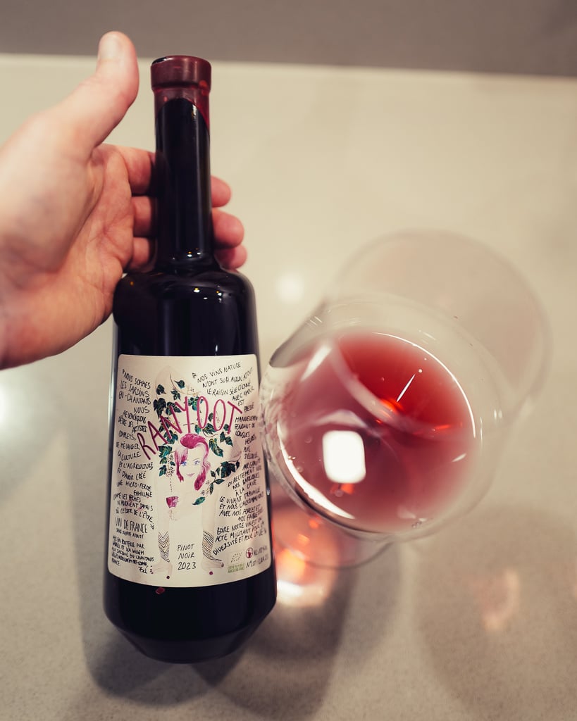 Hand houdt de donkere Ranyoot Pinot Noir met kunstzinnig etiket vast naast een glas helderrode wijn
