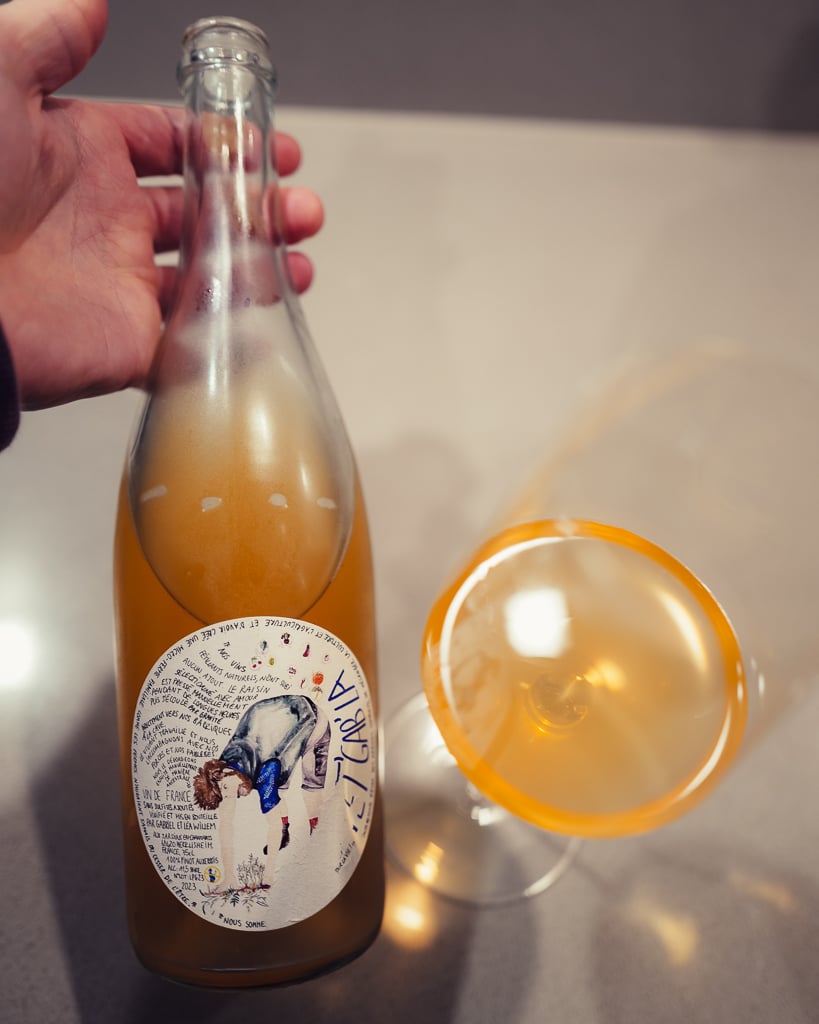 Hand houdt de Pet'Gab'La pétillant naturel vast naast een liggend glas met troebele oranje wijn