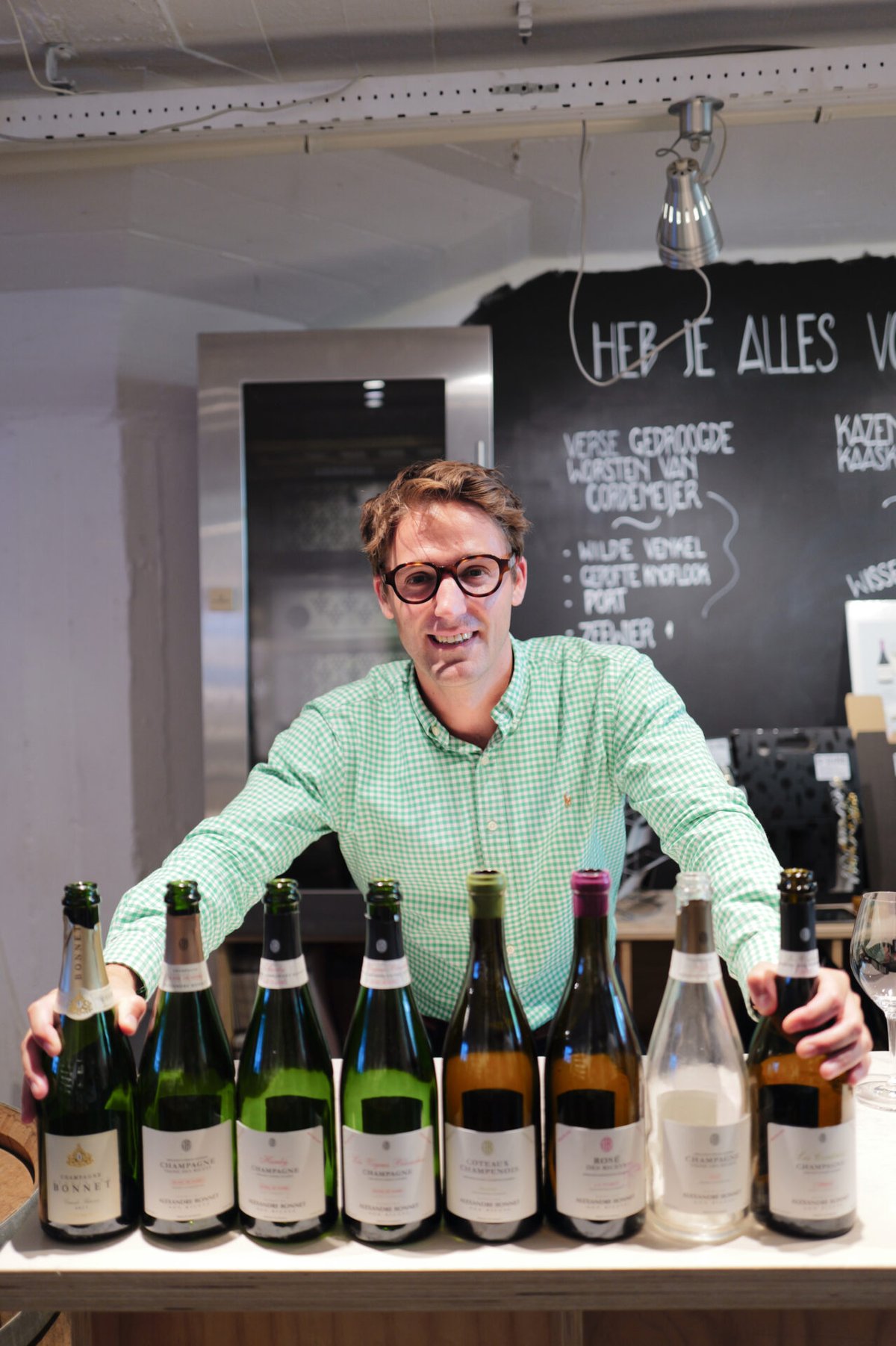 Man in groen geruit overhemd achter een rij van acht Champagne- en wijnflessen in een wijnwinkel