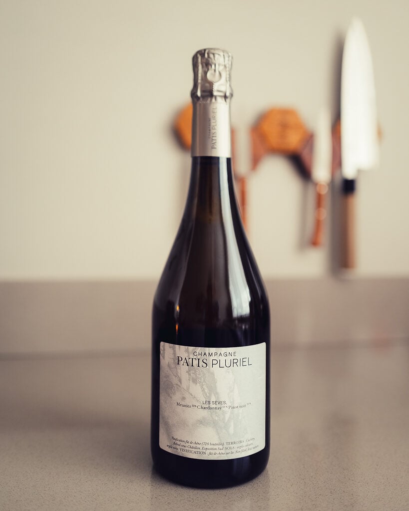 Fles Champagne Patis Pluriel Les Sèves rechtop op een aanrecht met een messenrek op de achtergrond