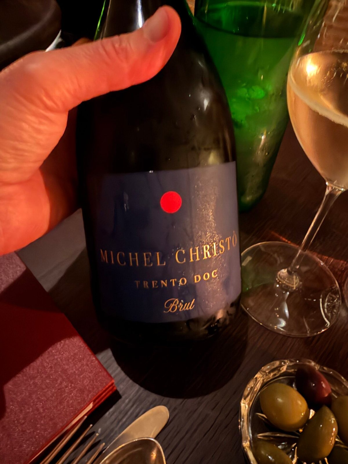 Hand houdt een fles Michel Christò Trento DOC Brut met blauw etiket vast aan een gedekte tafel met olijven