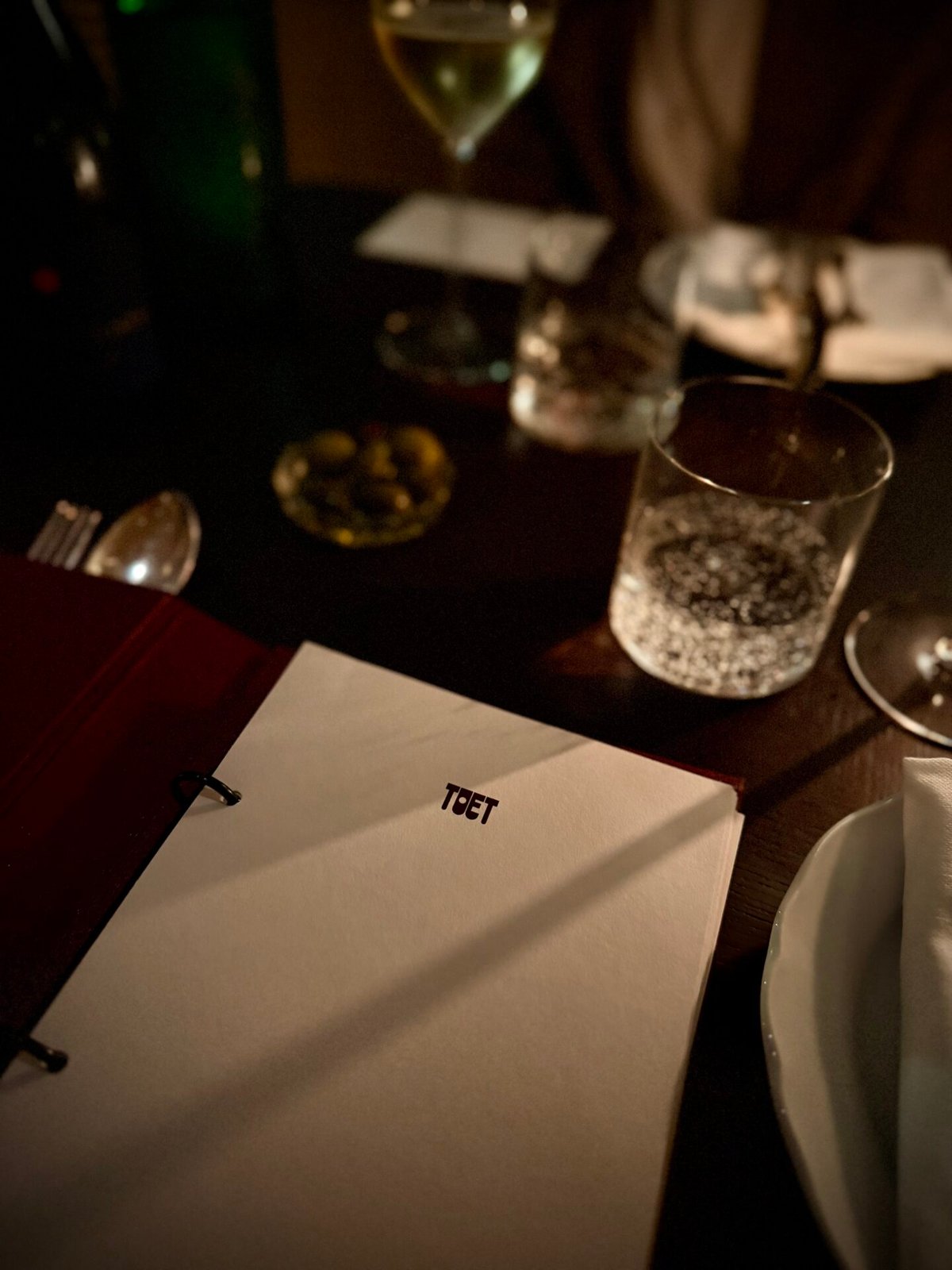 Menu met het logo TOET op een donkere gedekte tafel met waterglas en bestek in kaarslicht