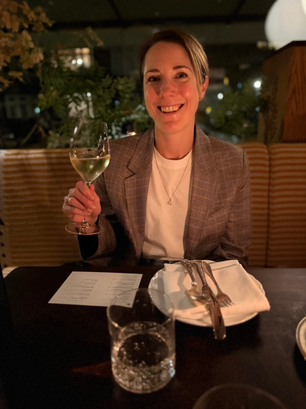 Lachende vrouw in geruit colbert heft een glas witte wijn aan een gedekte restauranttafel