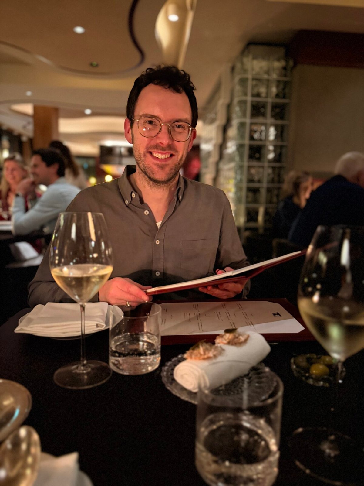 Lachende man met bril houdt een menu vast aan een restauranttafel met glazen witte wijn
