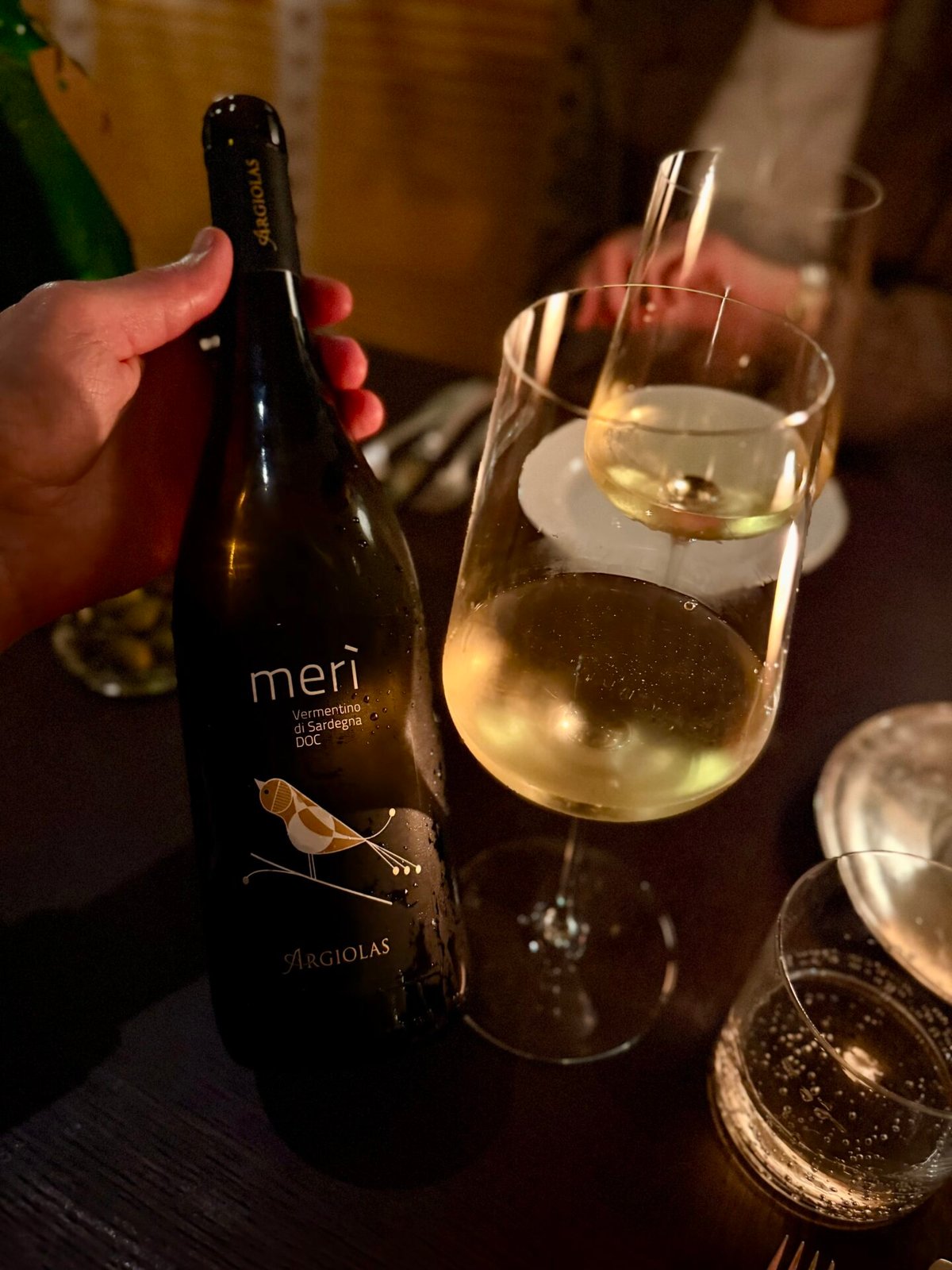 Hand houdt een fles Argiolas Merì Vermentino di Sardegna vast naast een glas witte wijn in kaarslicht