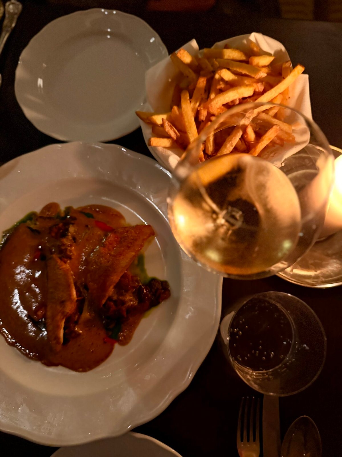Bord met visfilets in roodbruine saus naast een mandje friet en glazen wijn op een donkere tafel
