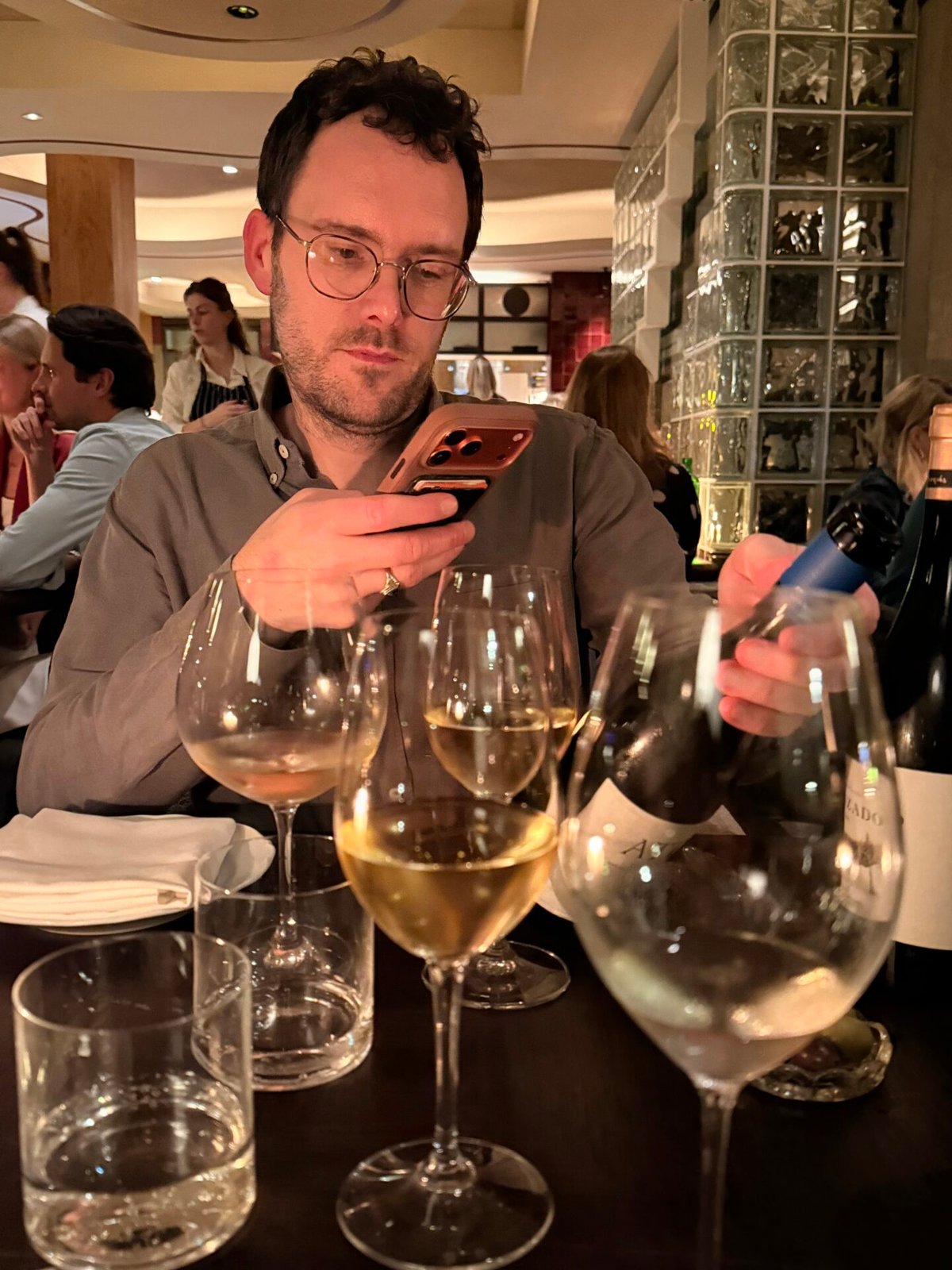 Man met bril aan een restauranttafel kijkt op zijn telefoon, glazen witte wijn op de voorgrond
