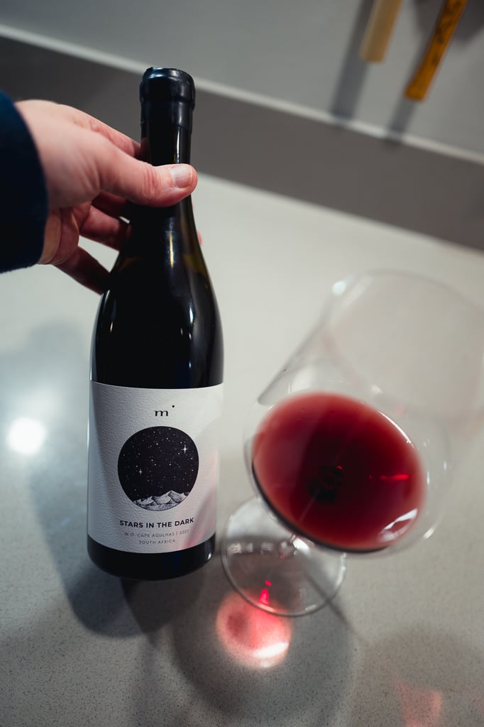 Hand houdt een fles Minimalist Stars in the Dark uit Cape Agulhas vast naast een glas rode wijn
