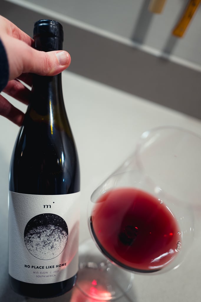 Hand houdt een fles Minimalist No Place Like Home Syrah vast naast een glas rode wijn