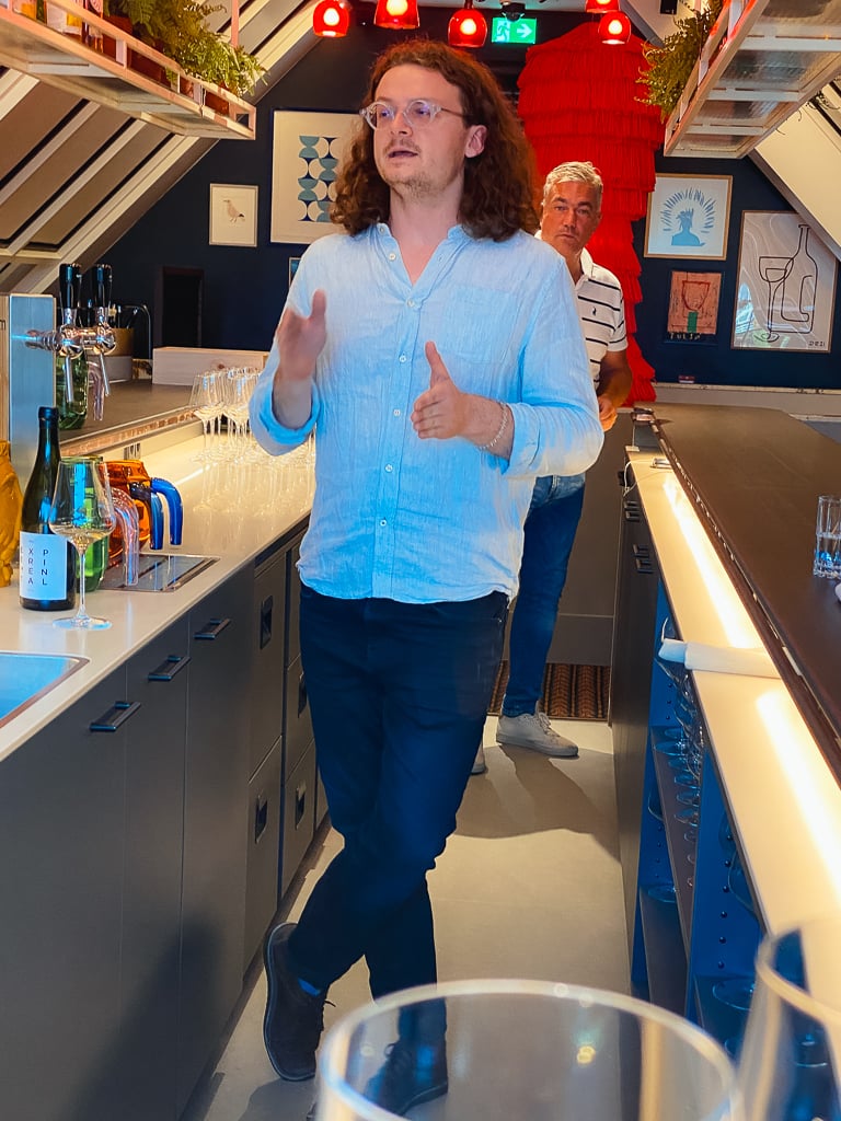 Wijnmaker in lichtblauw overhemd gebaart pratend in een bar terwijl een tweede man toekijkt
