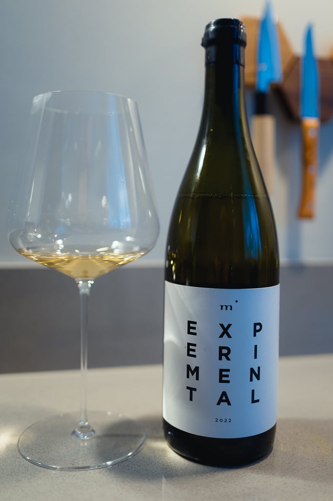 Fles Minimalist EXPERIMENTAL met wit etiket naast een glas witte wijn op een aanrecht