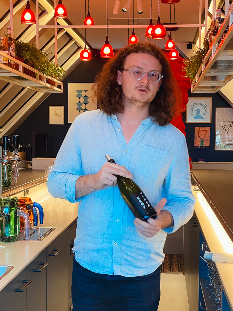 Wijnmaker met krullend haar in lichtblauw overhemd houdt een fles vast in een bar met rode hanglampen