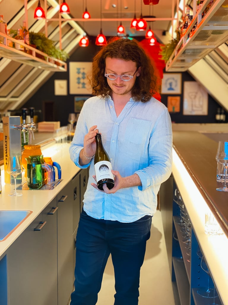 Wijnmaker in lichtblauw overhemd toont een fles met wit etiket in een bar met rode hanglampen
