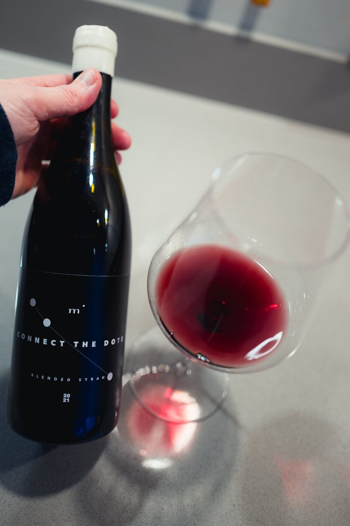 Hand houdt een fles Minimalist Connect the Dots Blended Syrah vast naast een glas rode wijn
