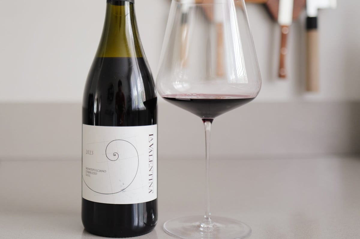 Fles La Valentina Montepulciano d'Abruzzo DOC naast een glas helderrode wijn op een lichte ondergrond
