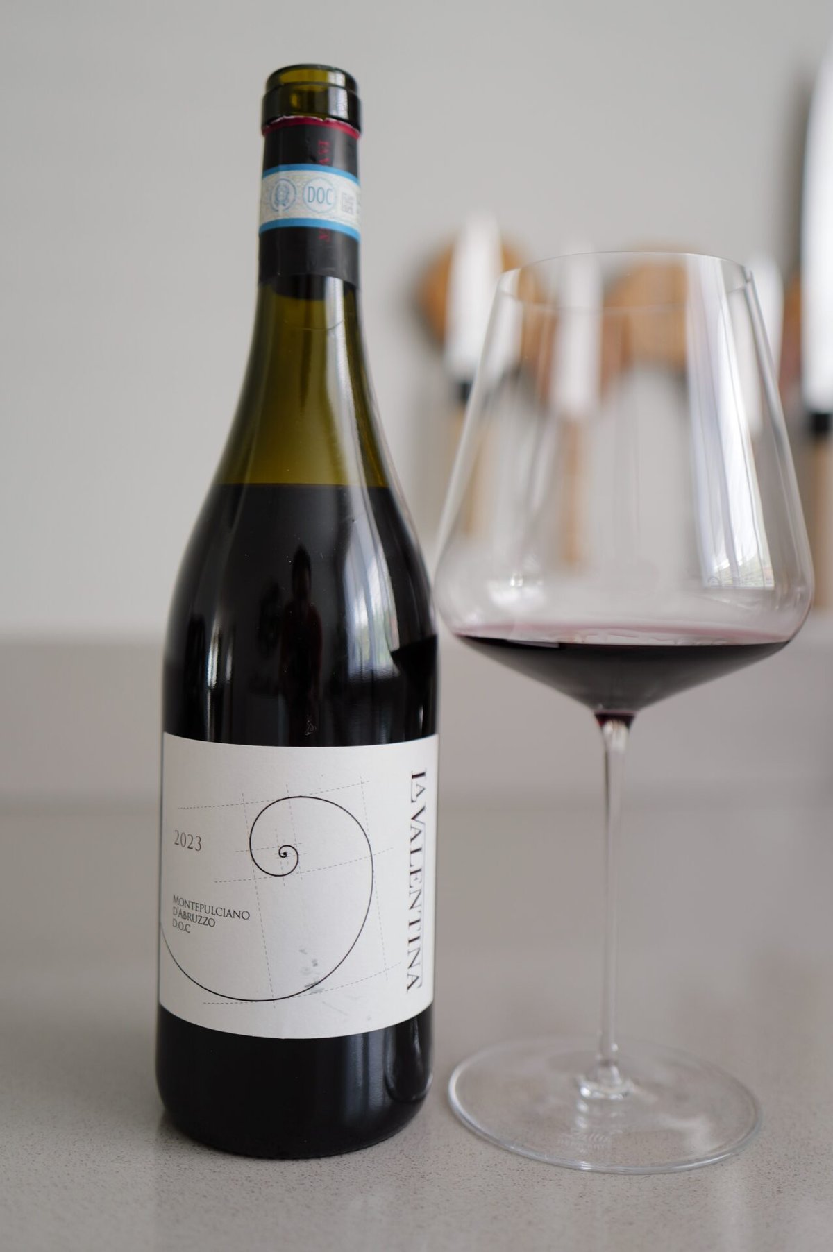 Fles La Valentina Montepulciano d'Abruzzo DOC met spiraal-etiket naast een glas rode wijn op een aanrecht