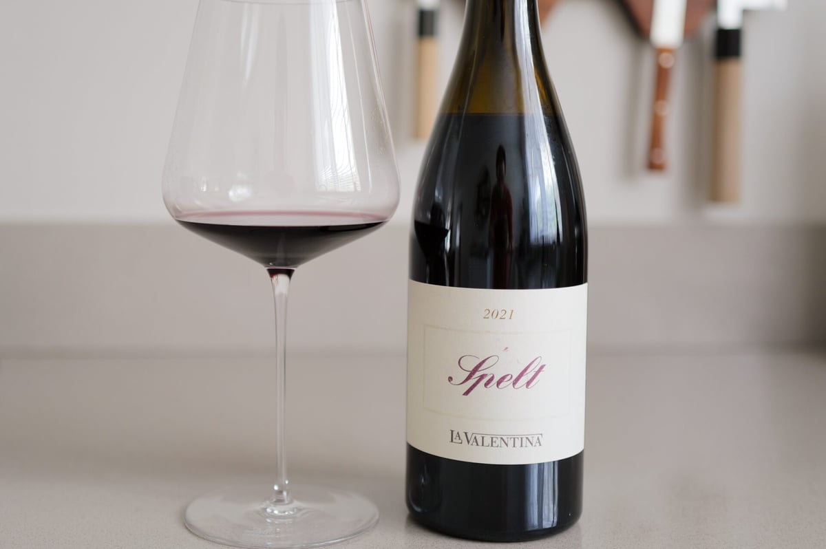 Glas rode wijn naast de fles La Valentina Spelt 2020 met sierlijk etiket op een lichte ondergrond