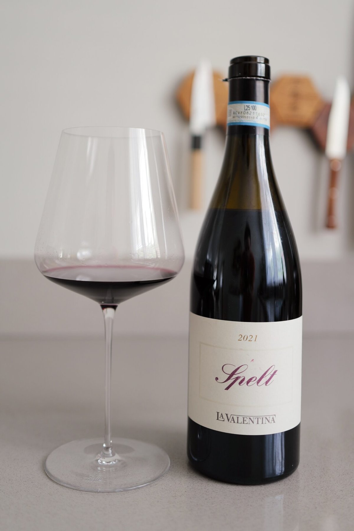 Fles La Valentina Spelt 2020 met sierlijk Spelt-etiket naast een glas rode wijn op een aanrecht
