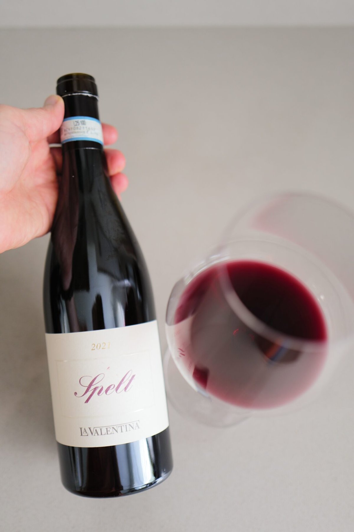Hand houdt de fles La Valentina Spelt 2020 vast naast een liggend glas rode wijn