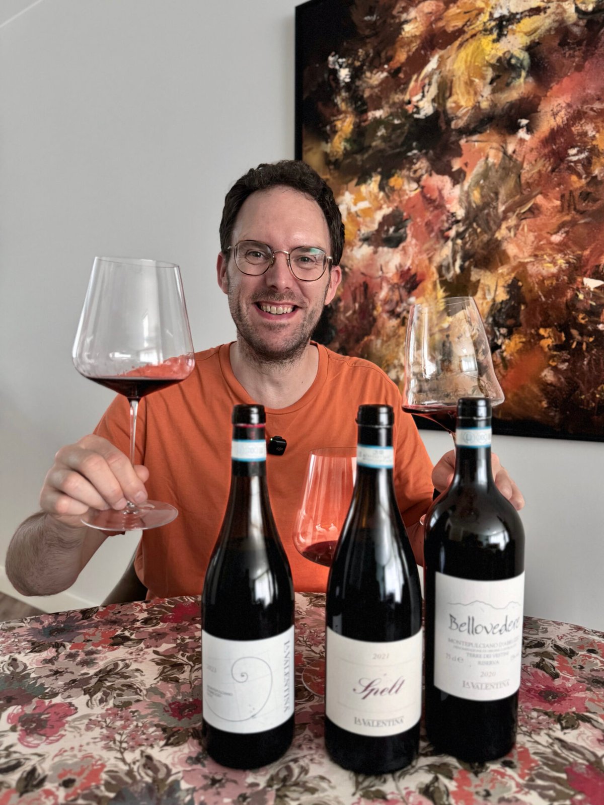 Lachende man in oranje shirt met wijnglas achter drie flessen rode La Valentina Montepulciano