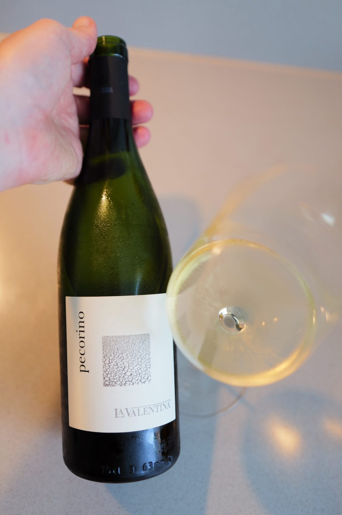 Hand houdt fles La Valentina Pecorino schuin naast liggend glas witte wijn