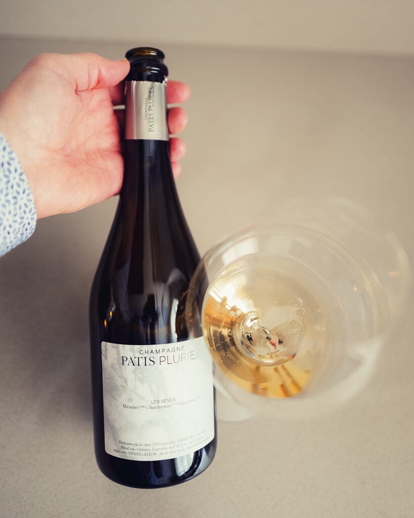 Hand houdt fles Champagne Patis Pluriel Les Sèves schuin naast liggend champagneglas