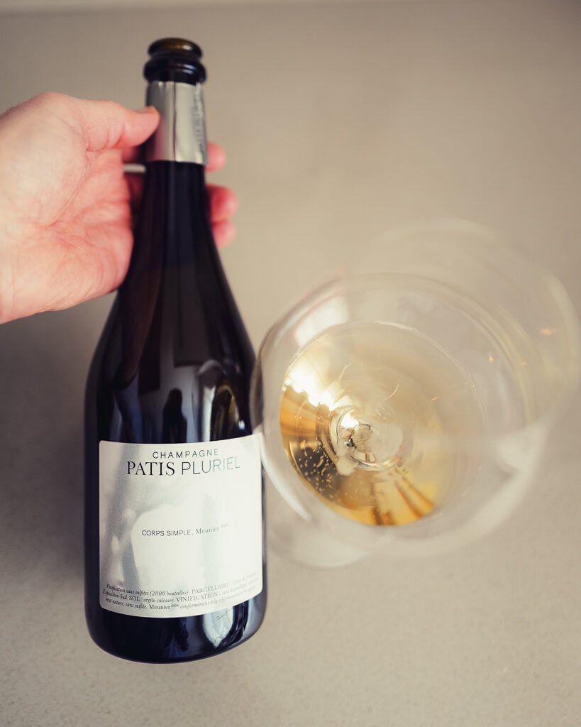 Hand houdt fles Champagne Patis Pluriel Corps Simple schuin naast liggend champagneglas