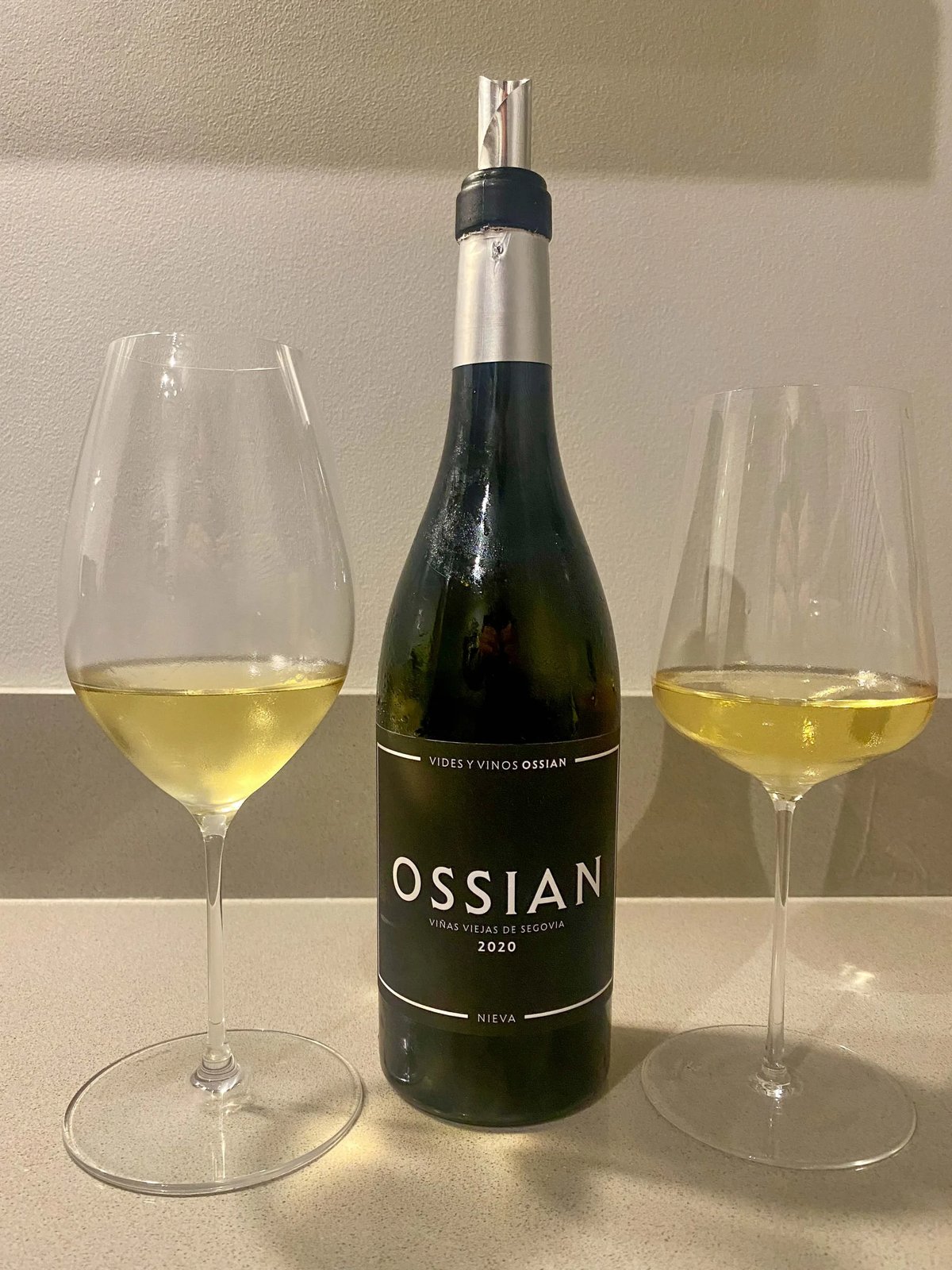 Fles Ossian witte wijn tussen twee glazen goudgele wijn tegen lichte muur